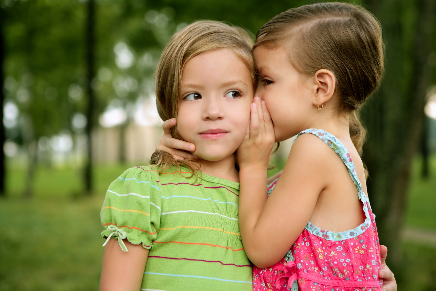 Two twin little sister girls whisper in the ear, outdoor on green park