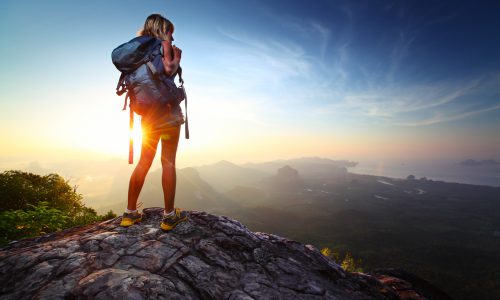 Young lady hiker standing with backpack on top of a mountain and enjoying sunrise